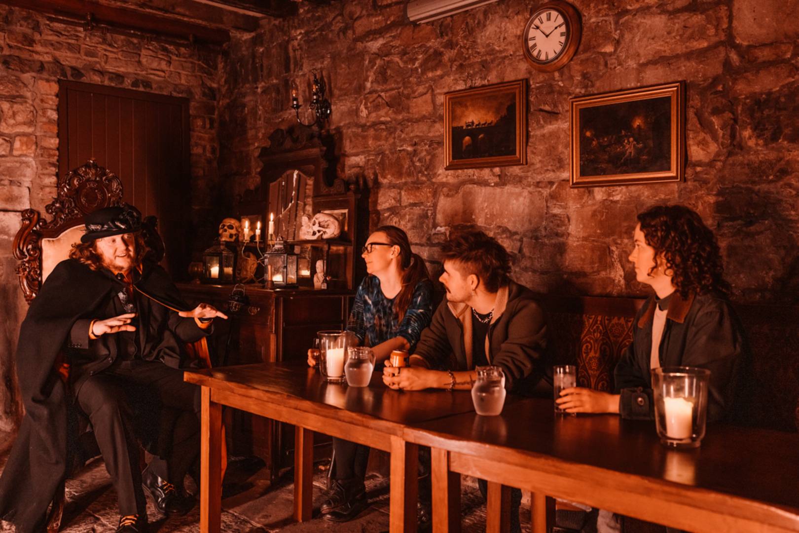 A Mercat Tours Storyteller sits at the end of a table in an underground cellar, telling ghost stories to a tour group sat with drinks.
